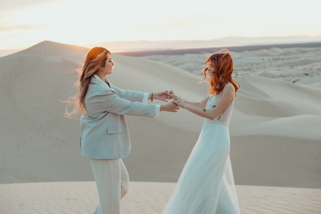 Two brides holding hands and dancing. Glamis Sand Dunes in California provide the backdrop. 