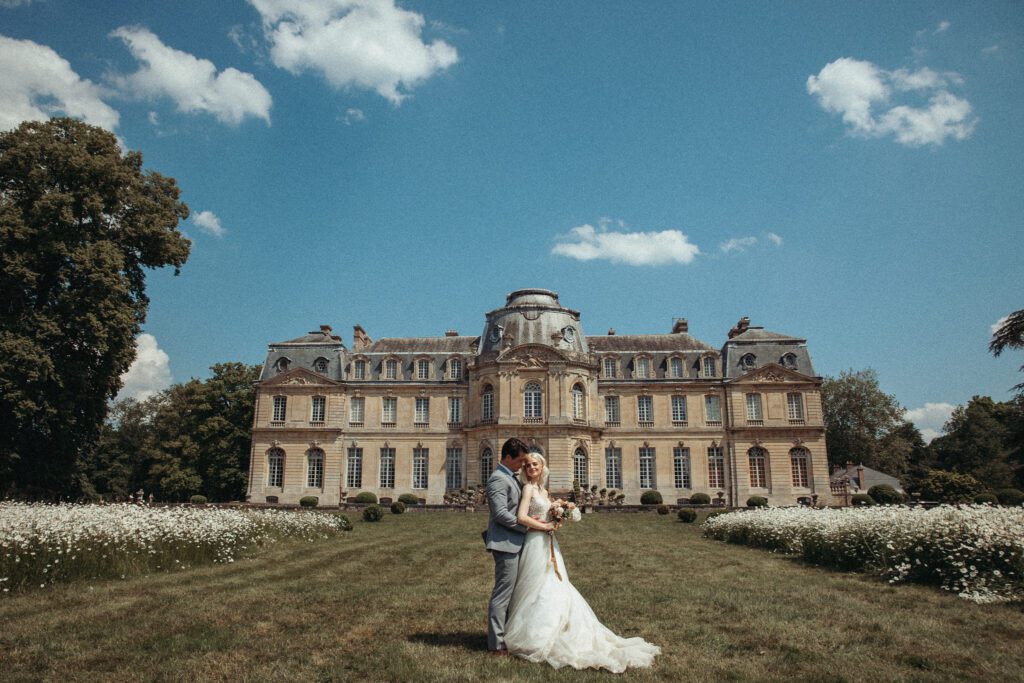 Bride and groom standing in front of  chateau de champlatreux in France surrounded by wildflowers on a sunny day. 