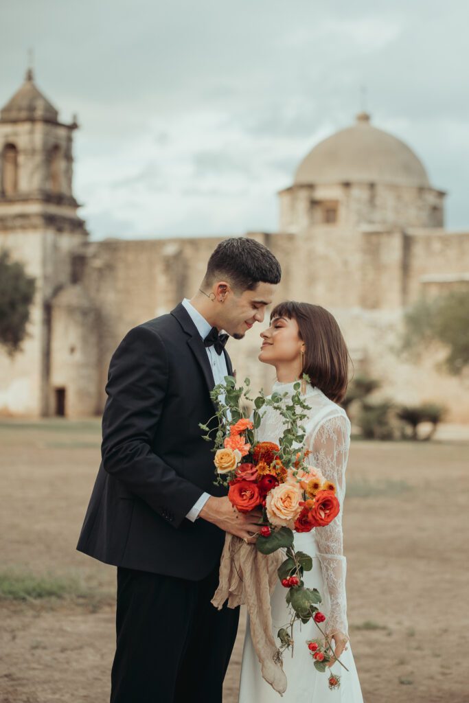 Bride and groom in front of The Missions in San Antonio about to share a kiss. The bride is holding a large, colorful bouquet with bright red flowers. 