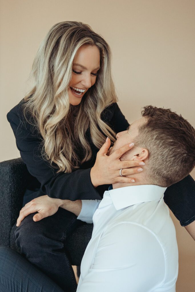 Bride sitting in chair while groom is sitting on the ground. Bride is holding the groom's face and looking into his eyes and smiling.